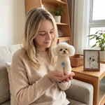A woman in a beige sweater sits on a sofa in a bright living room, holding a small fluffy white dog dressed in a beige sweater with a blue circular tag. A framed photo of a similar dog sits on the wooden table beside her.